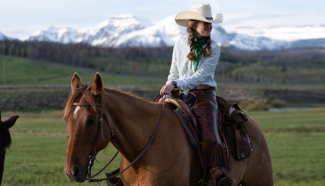 A woman wearing a cowboy hat and chaps riding on horseback in the Wyoming wilderness.