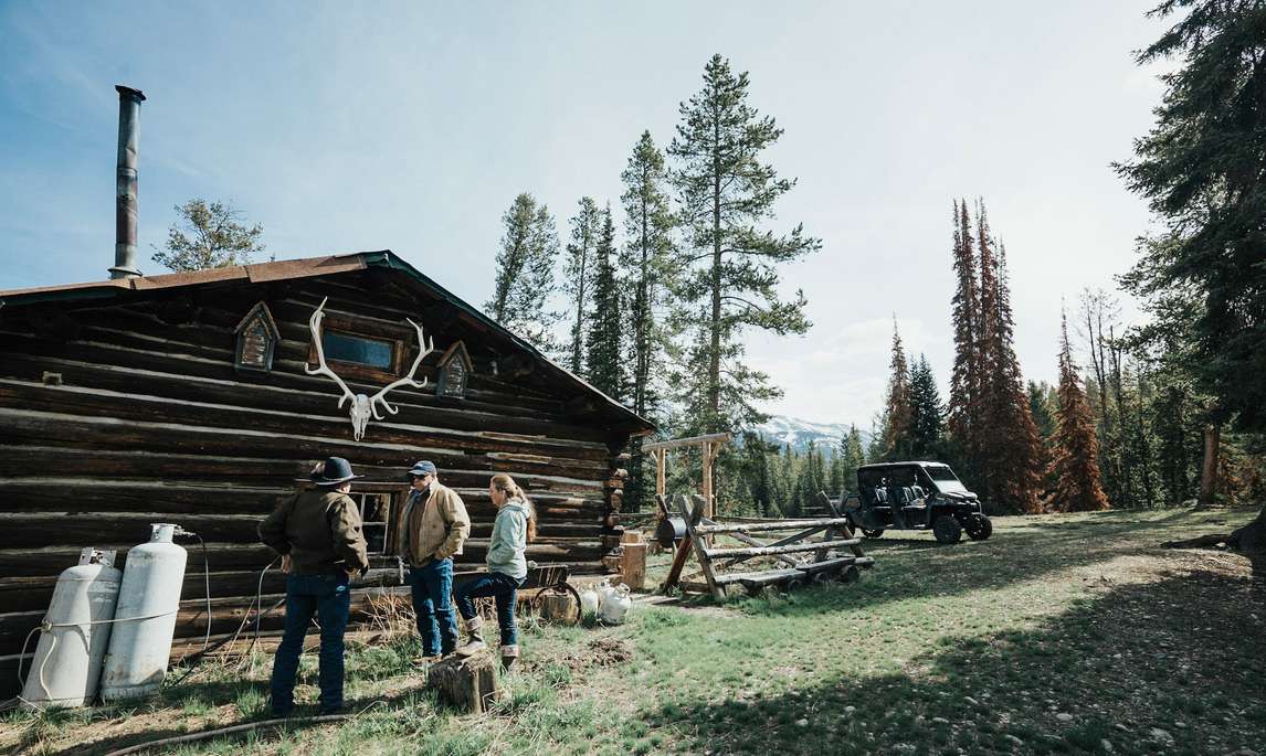 Three people standing near the historic Craig Cabin at the Jack Creek Camp in Wyoming.